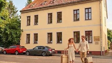 Historisches Gebäude mit gelber Fassade und roten Dachziegeln, an einer Straße gelegen.
