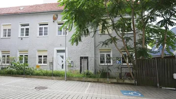 Graues Mehrfamilienhaus mit gepflastertem Parkplatz, grünen Bäumen und Rollläden an den Fenstern.