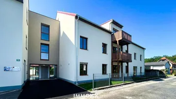 Modernes Mehrfamilienhaus mit heller Fassade, Balkonen und gepflegter Außenanlage unter blauem Himmel.