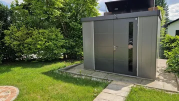 Moderner Gartenschuppen mit Solarpaneelen auf dem Dach, umgeben von Rasen und Sträuchern.