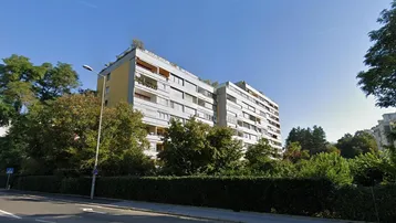 Mehrstöckiges Wohngebäude mit Balkonen und Flachdach, umgeben von Bäumen und einer Hecke an einer Straße.