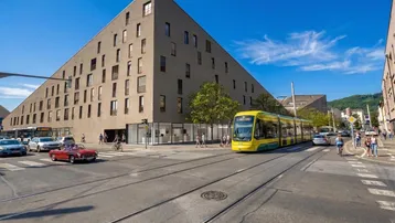 Modernes, eckiges Gebäude an einer belebten Kreuzung mit Straßenbahn und Passanten in urbaner Umgebung.