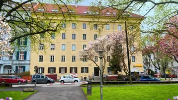 Gelbes Mehrfamilienhaus mit grüner Parkanlage und blühenden Bäumen im Frühling.