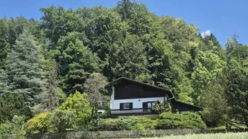 Traditionelles Haus mit Satteldach, umgeben von üppiger grüner Natur und Wald am Hang.