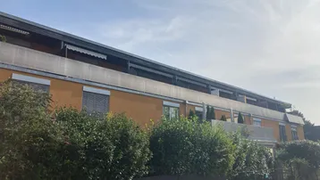 Gebäudefassade mit mehreren Balkonen und grünen Hecken im Vordergrund.