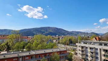 Panoramablick über die Stadt mit Wohngebäuden und grünen Hügeln im Hintergrund unter blauem Himmel.