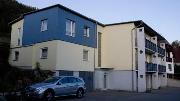 Gebäudeansicht mit blauer Holzverkleidung und gelber Fassade, ein Auto parkt davor.