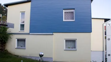 Seitenansicht des Gebäudes mit blauer Verkleidung und gelber Fassade, umgeben von Grün.