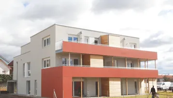 Modernes Mehrfamilienhaus im Bau mit roten Akzenten und Balkonen unter bewölktem Himmel.
