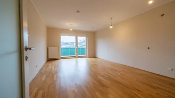 Helles, leeres Wohnzimmer mit Parkettboden und Zugang zu einem Balkon.