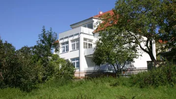 Weißes Gebäude mit Balkonen, umgeben von Bäumen und grüner Landschaft.