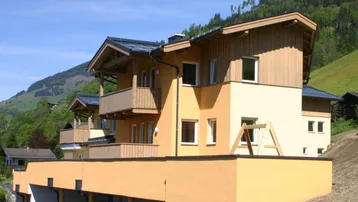 Modernes Mehrfamilienhaus mit Holzverkleidung, Balkonen und Carports vor alpiner Bergkulisse.
