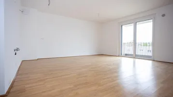 Helles, leeres Wohnzimmer mit Parkettboden und Zugang zum Balkon.