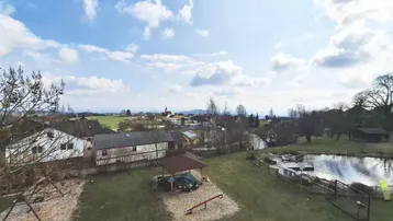 Weitläufiger Blick über eine ländliche Umgebung mit Häusern, einem Teich und einem Spielplatz unter blauem Himmel.