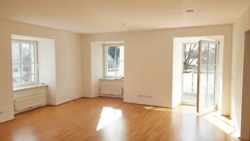 Helles Zimmer mit Parkettboden, weißen Wänden und Zugang zu einem Balkon.