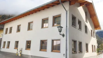 Traditionelles Gebäude mit weißer Fassade, Holzbalken und Satteldach in alpiner Umgebung.