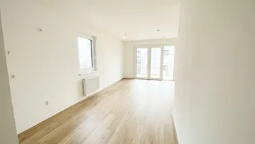 Helles, leeres Zimmer mit Holzfußboden und Zugang zu einem Balkon in einem Neubau.