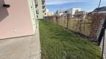 Seitlicher Gartenbereich mit gepflasterter Terrasse und Blick auf die moderne Wohnanlage.