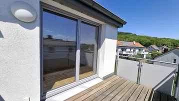Heller Balkon mit Holzboden und Blick auf die grüne Umgebung, zugänglich durch eine große Schiebetür.