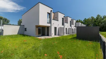 Moderne Reihenhäuser mit weißer Fassade und direktem Zugang zu grünen Gärten unter blauem Himmel.