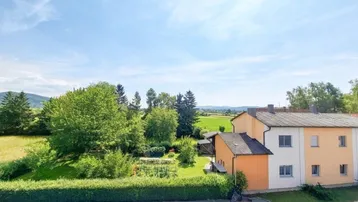 Ländliche Umgebung mit grünen Feldern, Bäumen und einem Wohnhaus unter blauem Himmel.