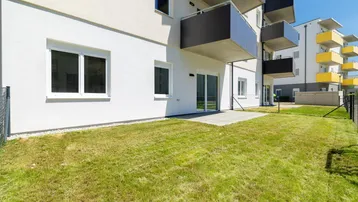Gepflasterte Terrasse mit Rasenfläche vor einem modernen Wohngebäude bei sonnigem Wetter.