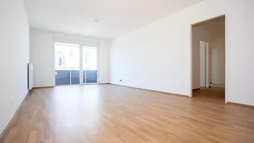 Helles, leeres Wohnzimmer mit Laminatboden, weißen Wänden und Zugang zu einem Balkon.