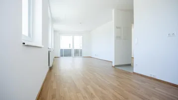 Helles, leeres Wohnzimmer mit Holzboden, weißen Wänden und Zugang zu einem Balkon.