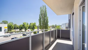 Heller Balkon mit Blick auf einen Parkplatz und umliegende Geschäfte unter blauem Himmel.