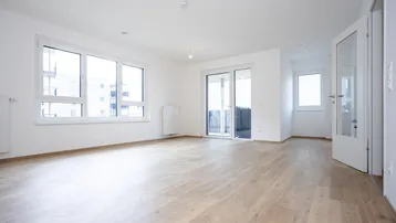 Helles, leeres Wohnzimmer mit Laminatboden, großen Fenstern und Zugang zum Balkon.