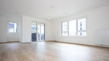 Geräumiges Wohnzimmer mit hellem Laminatboden, großen Fenstern und Zugang zu einem Balkon.