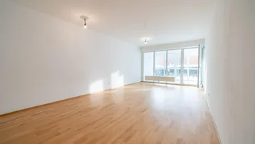 Helles, leeres Wohnzimmer mit Parkettboden und großen Fenstern, die viel Tageslicht hereinlassen.