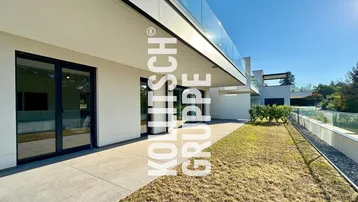 Moderne Terrasse mit Fliesenboden und Rasenfläche, umgeben von einer Glasbrüstung unter blauem Himmel.