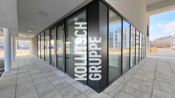Moderne Geschäftsfassade mit großen Glasfenstern und gepflastertem Außenbereich, Teil eines Neubaus.