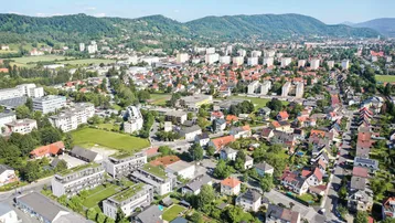 Weitläufige Luftaufnahme einer Wohngegend mit grünen Hügeln und städtischer Bebauung im Hintergrund.