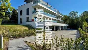 Modernes Mehrfamilienhaus mit Balkonen, Glasgeländern und Tiefgaragenzufahrt in sonniger Umgebung.