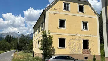 Historisches Gebäude mit gelber Fassade und sichtbaren Schäden, umgeben von Natur und Bergen unter blauem Himmel.