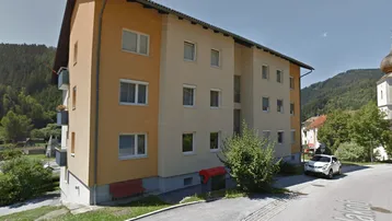 Mehrfamilienhaus mit schräger Dachkonstruktion, orangefarbener Fassade und umliegender Naturkulisse.
