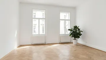 Heller Raum mit Fischgrätparkett und zwei großen Fenstern, die viel Tageslicht hereinlassen.