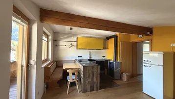 Helle offene Wohnküche mit Holzbalken, gelber Wand und rustikalem Essbereich mit Bergblick.