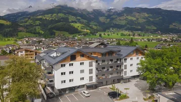 Modernes Mehrfamilienhaus mit Balkonen und Bergblick, eingebettet in eine grüne Landschaft mit umliegenden Häusern.