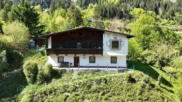 Traditionelles Haus im alpinen Stil mit Holzbalkon und bemalter Fassade, umgeben von grünen Hügeln.