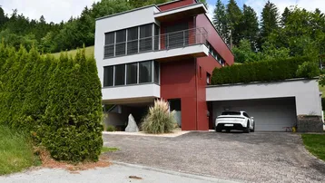 Modernes Haus mit roter und grauer Fassade, umgeben von grünen Hügeln und Bäumen.