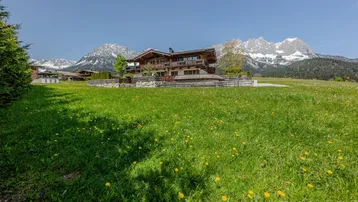 Großes Holzchalet in einer grünen Wiese mit Alpenpanorama und schneebedeckten Bergen im Hintergrund.