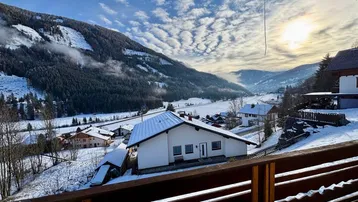 Winterlandschaft mit Blick auf schneebedeckte Berge und ein Haus im Tal unter einem bewölkten Himmel.