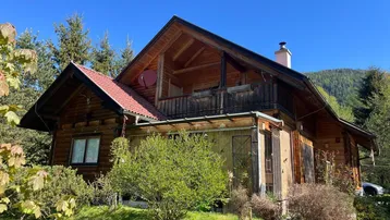 Charmantes Holzhaus mit rotem Dach und Balkon, umgeben von grüner Natur und Bergen.