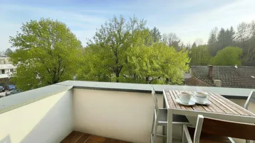 Heller Balkon mit Holzbodenfliesen, kleinem Tisch und Blick ins Grüne.