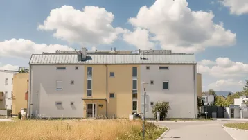 Mehrfamilienhaus mit moderner Fassade und umliegender Grünfläche unter blauem Himmel.