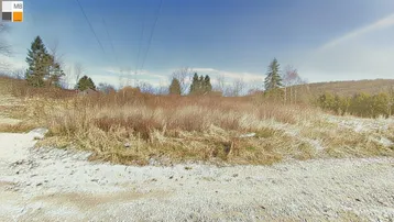 Weites Grundstück mit trockener Vegetation und leichtem Schneefall, umgeben von Bäumen und Hügeln unter blauem Himmel.