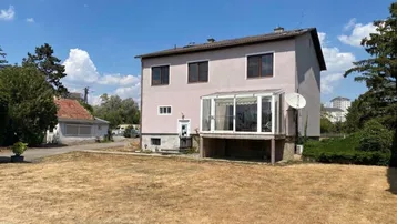 Hausfassade mit Wintergarten und großem, trockenem Gartenbereich unter blauem Himmel.